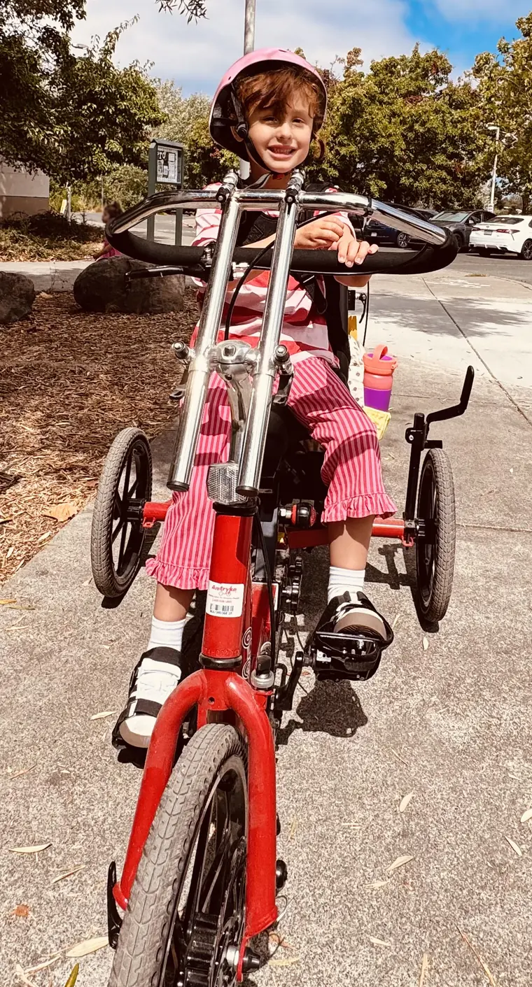 A young rider seated frontally on a red adaptive tricycle outdoors.