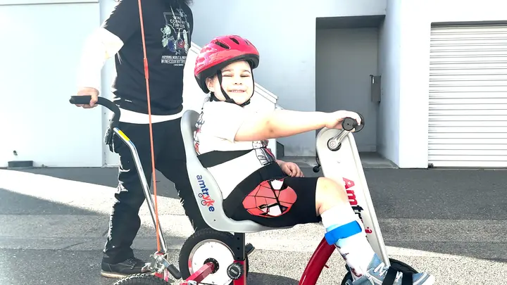 Lukas riding his earlier adaptive Tryke with support nearby.